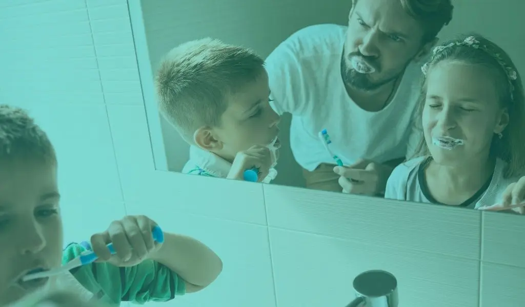 Genitore insegna ai bambini come lavarsi i denti davanti allo specchio, promuovendo una corretta igiene orale